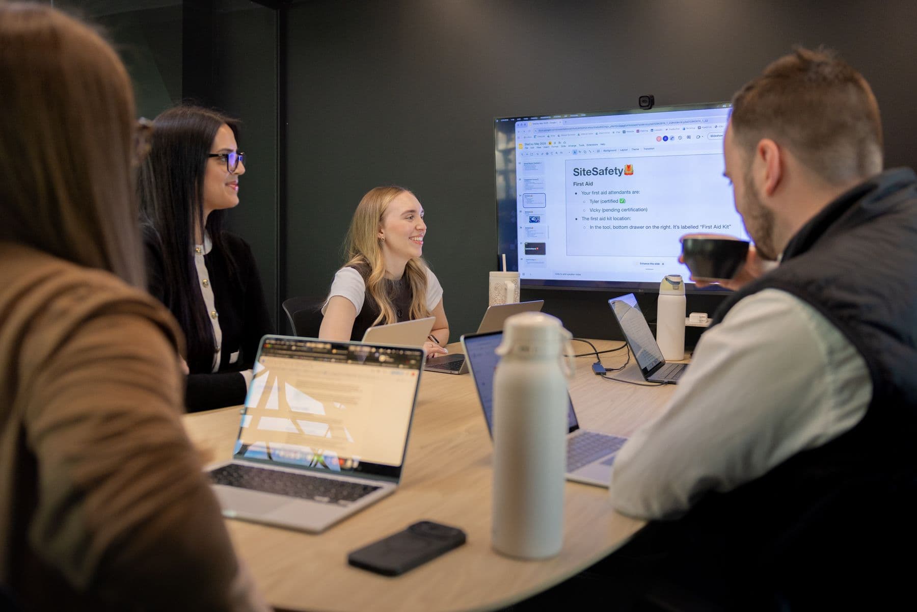 People sit at boardroom table with laptops, looking at a presentation on screen.