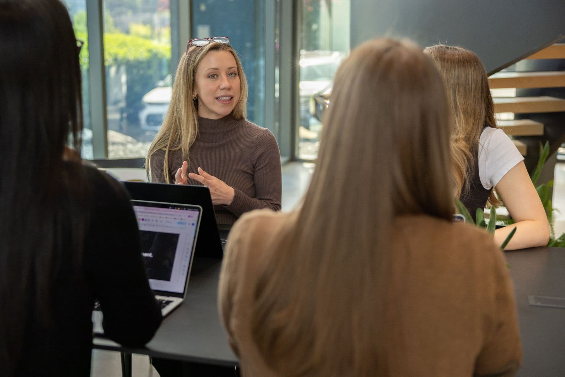 A person is taking to a table of people. They all have laptops and are in an office environment.
