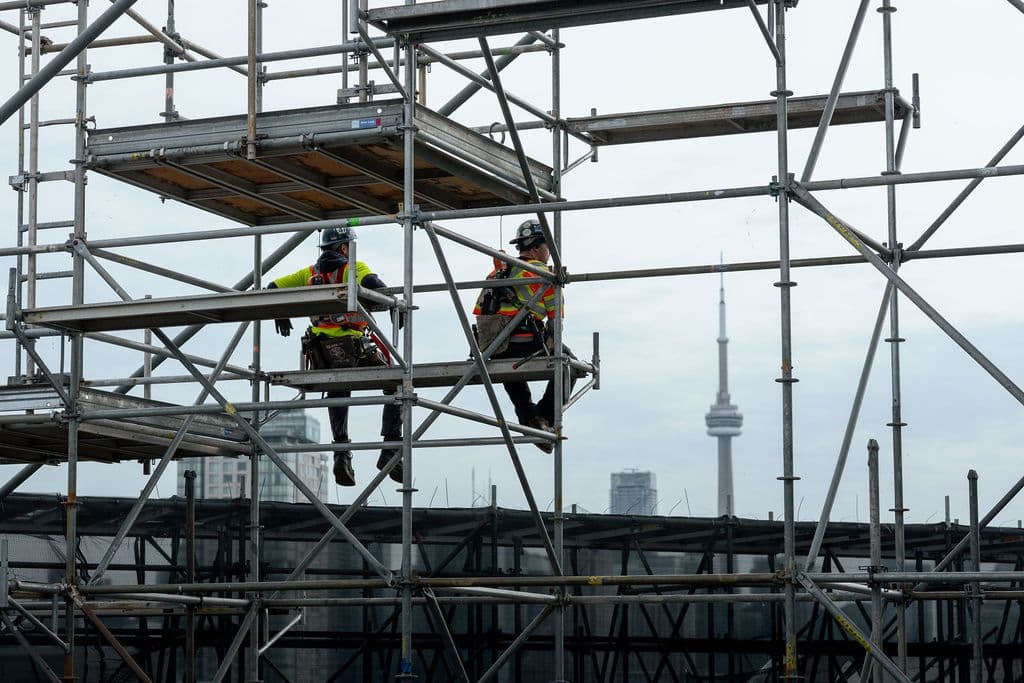 Two people in hard hats and high vis sit in a steel contraption at a construction site, looking at the view of the CN Tower in Toronto.