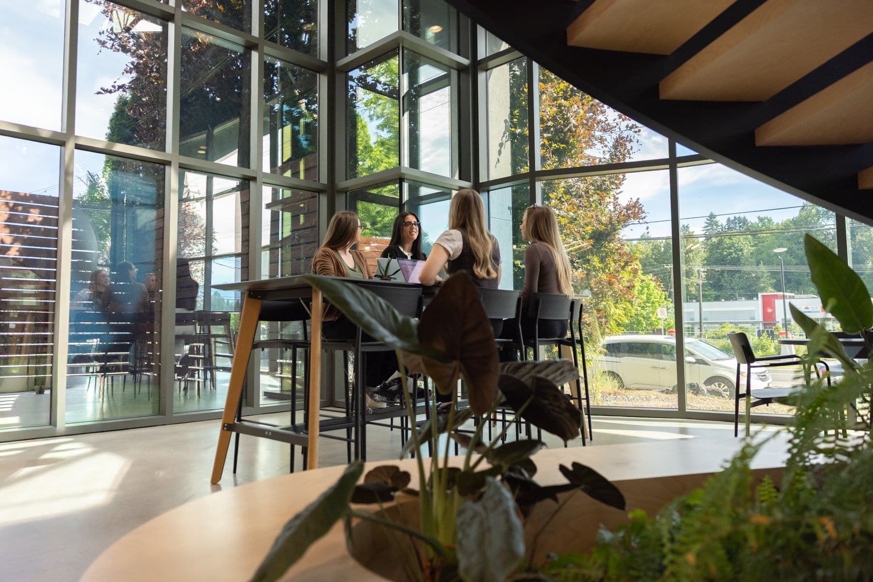 Four people site at a table with laptops, talking to one another, in an open office space with windows in the background.