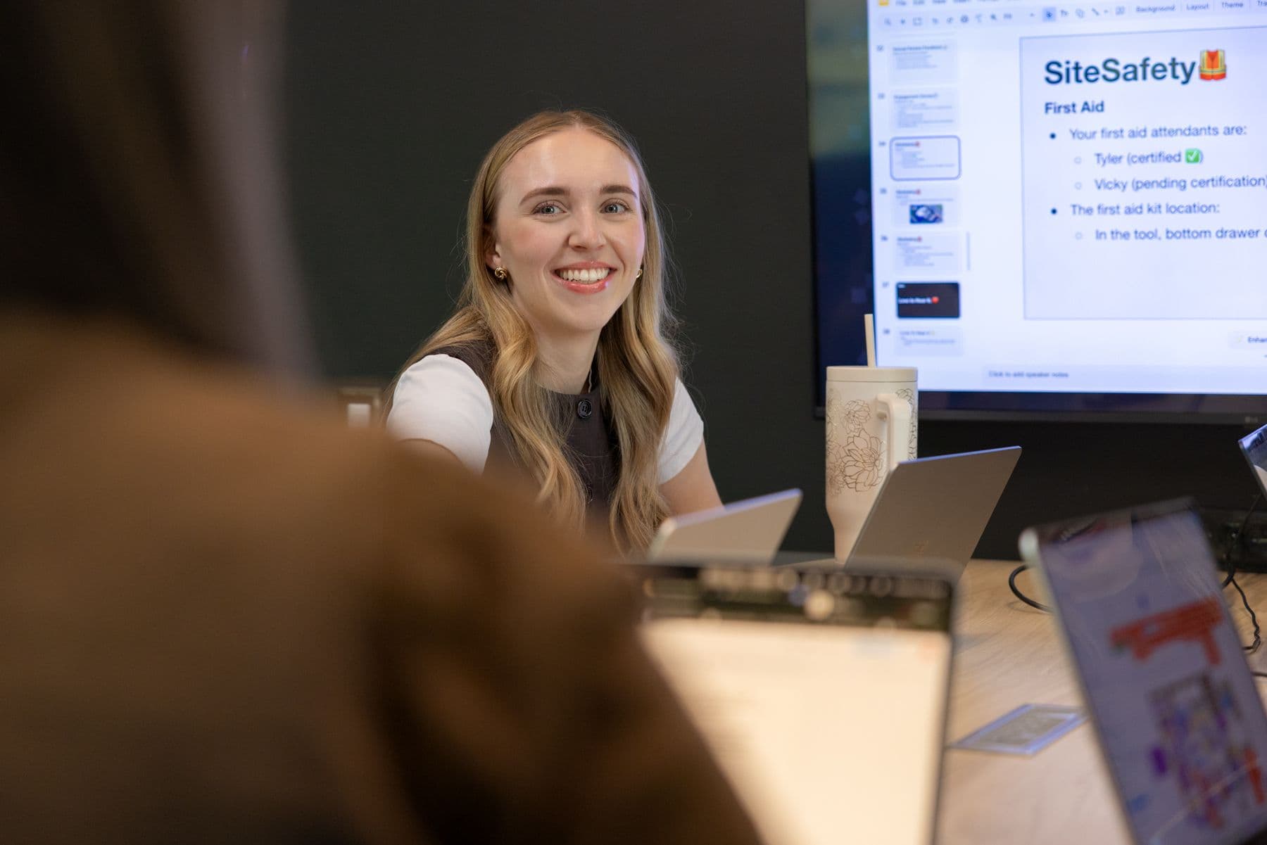 A woman smiling at a person across the table. They have laptops in front of them and a presentation in the background.